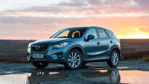 藍色Mazda CX-5 SUV停泊在濕碎石路上，水坑中映出日落倒影。 — 馬自達 壁紙