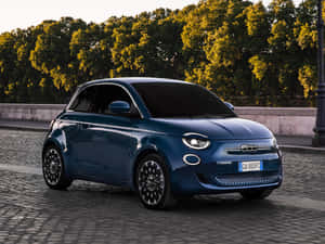 藍色電動Fiat 500e在鵝卵石街道上，有樹木和夕陽。 — 飛雅特 壁紙