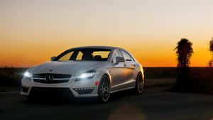 銀色 Mercedes-Benz CLS63 AMG 在日落時停放，車頭燈發光。 — 日落 壁紙