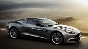銀色Aston Martin跑車行駛在山路上，背景是霧氣瀰漫的山谷和夕陽餘暉。 — 奧斯頓馬丁 壁紙