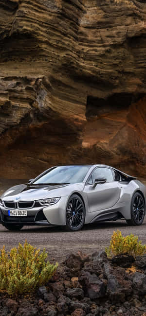 銀色BMW i8跑車停放在岩石懸崖下，伴有沙漠灌木。 — Pixel 8 Pro 壁紙