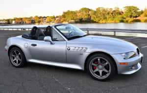 銀色BMW Z8敞篷車停泊在水邊附近，背景有樹木和日落。 — Steam Deck OLED 壁紙