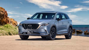 銀色Mazda CX-90 SUV停泊在海洋旁邊，背景有岩石峭壁和藍天。 — 馬自達 壁紙