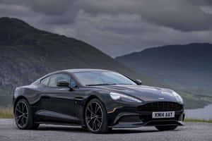 黑色Aston Martin跑車停泊在碎石上，靠近山脈，天空多雲。 — Surface Laptop 壁紙