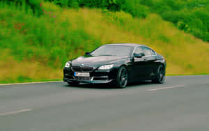 黑色BMW 6系列在風景優美的道路上行駛，穿過綠色和黃色的草地。 — Steam Deck 壁紙