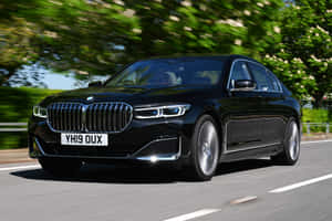 黑色BMW 7系列轎車在陽光明媚的道路上行駛，周圍有綠色樹木。 — Surface Pro 壁紙