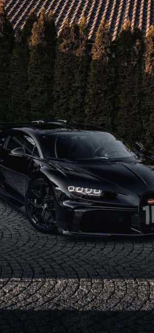 黑色Bugatti Chiron停泊在鵝卵石車道上，伴隨著常綠樹和夕陽。 — iPhone 18 壁紙