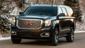 黑色GMC Yukon SUV在雪路上行駛，兩旁是冬日禿樹。 — Gmc 壁紙