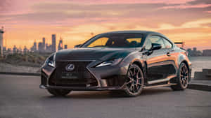 日落時分，黑色Lexus RCF Track Edition停在海濱，背後是城市天際線。 — Studio Display 壁紙