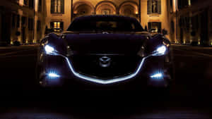 黑色Mazda跑車在夜間停在燈光明亮的建築前，頭燈發光。 — 馬自達 壁紙
