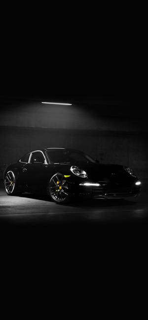 黑色保時捷911在光線昏暗的地下車庫中，有單一頭頂燈。 — iPhone 15 Pro 壁紙