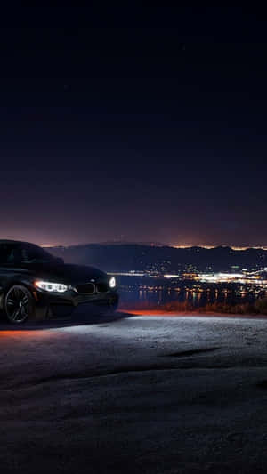 黑色跑車停在山坡上，俯瞰夜間的城市燈光。 — BMW iPhone 壁紙