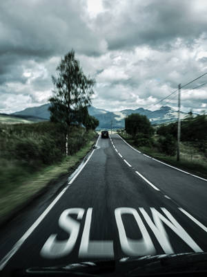 一輛汽車在塗有「SLOW」的道路上行駛，背景是陰雲密布的天空下的山巒。 — 汽車 iPhone 壁紙