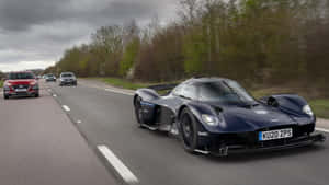 深藍色 Aston Martin Valkyrie 在潮濕的道路上與其他車輛和樹木一起疾馳。 — 奧斯頓馬丁 壁紙