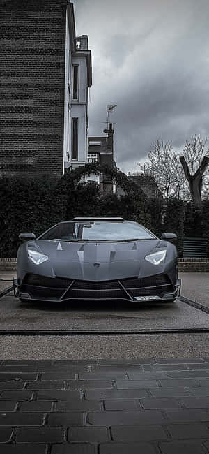 一輛灰色的藍寶堅尼 Aventador 停泊在濕漉漉的鵝卵石街道上，天空多雲。 — iPhone 17 壁紙