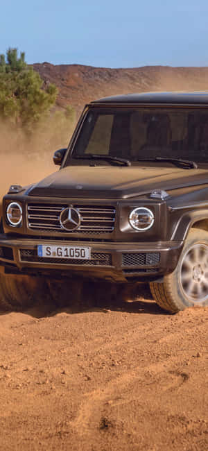 黑色 Mercedes G-Class 在塵土飛揚的沙漠地形中行駛，有丘陵和樹木。 — Galaxy S25 FE 壁紙