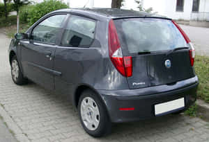 深灰色Fiat Punto停在磚砌車道上，附近有建築物和樹木。 — 飛雅特 壁紙