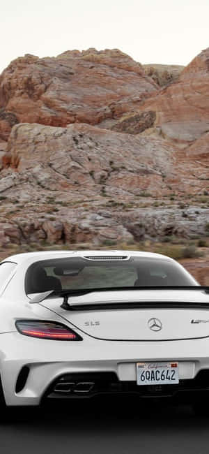 白色 Mercedes SLS AMG 在日落時分疾馳穿過紅色岩石沙漠峽谷。 — iPhone 15 Plus 壁紙