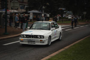 白色BMW M3拉力賽車在潮濕的城市街道上行駛，有觀眾觀看。 — 白色 iPhone 壁紙