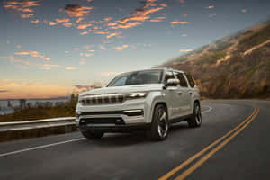 白色Jeep Grand Wagoneer在日落時分於城市附近的蜿蜒山路上行駛。 — Surface Pro 壁紙