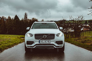 白色Volvo XC90 SUV在潮濕的鄉村道路上，多雲天空和綠色田野。 — 富豪 壁紙