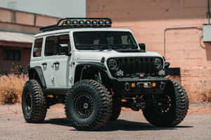 白色升高的Jeep Wrangler，配備大輪胎，停泊在沙漠工業建築附近。 — MacBook Air M2 壁紙