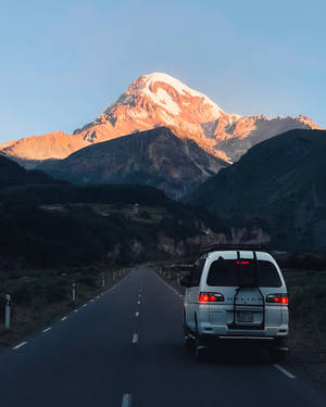 白色廂型車在山路上接近夕陽下的雪山頂峰。