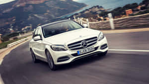白色賓士轎車在蜿蜒的沿海道路上行駛，背景有山脈和海洋。 — Mercedes Benz C300 壁紙