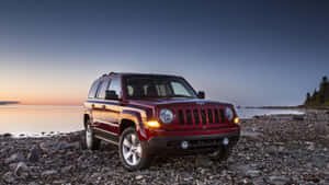 紅色Jeep Patriot在日落時停泊在岩石湖岸，靠近平靜的水面。 — 吉普 壁紙