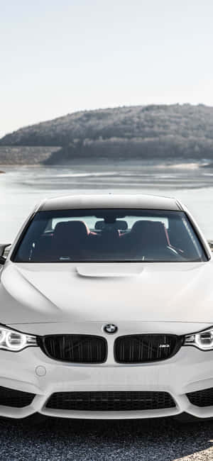 白色BMW M4停泊在湖邊，後面有樹木繁茂的山丘。 — Pixel 7a 壁紙