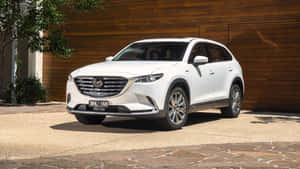 白色馬自達CX-90 SUV停放在石頭車道上，靠近木牆。 — 馬自達 壁紙