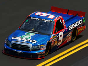 帶有 GEICO 標誌的藍色和紅色 NASCAR 賽車卡車在賽道上飛馳。 — 汽車 壁紙