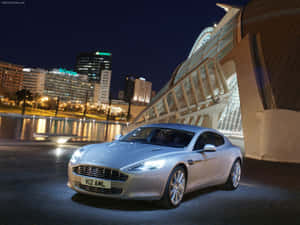 銀色Aston Martin夜間停放在現代玻璃橋旁，城市燈光倒映在水中。 — 奧斯頓馬丁 壁紙