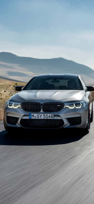 銀色BMW M5在沙漠公路上疾駛，背景有山脈，日落時分。 — Redmi Note 14 Pro 壁紙