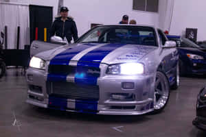 銀色和藍色的日產Skyline R34，帶有賽車條紋，在車展上展出。 — 湯姆克魯斯 壁紙
