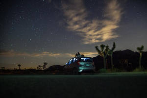 兩個人坐在銀色汽車車頂上，背景是繁星點點的沙漠夜空和約書亞樹。 — 暮光 壁紙