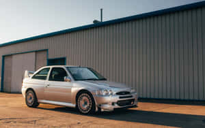 日落時分，銀色福特 Escort RS Cosworth 停在工業建築外。 — MacBook Pro 13 壁紙
