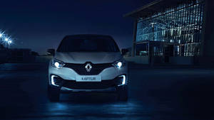 銀色雷諾Captur SUV在夜晚被車頭燈照亮，停在一棟現代玻璃建築前。 — 雷諾 壁紙