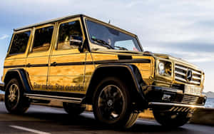 金色 Mercedes G-Class SUV 在海岸公路上行駛，背景是海洋。 — 金色動態 壁紙