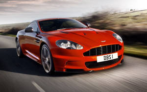 紅色Aston Martin DBS在日落時分在風景優美的道路上疾馳。 — 奧斯頓馬丁 壁紙