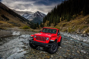 紅色Jeep Rubicon穿越岩石山間溪流，背景有白雪覆蓋的山峰。 — 山景 壁紙