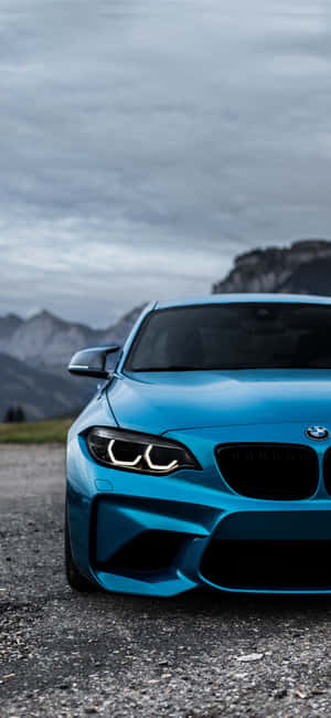 藍色BMW跑車停在碎石上，靠近山脈，天空多雲 — iPhone 15 Pro Max 壁紙