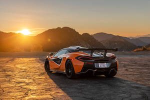 橙色McLaren跑車停在裂開的路面上，日落時分，背後有山脈。 — 跑車 壁紙