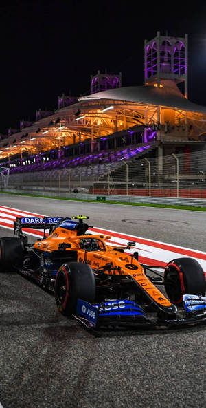 夜間賽道上橙色和藍色的F1賽車，看台燈光璀璨。 — 賽車 壁紙