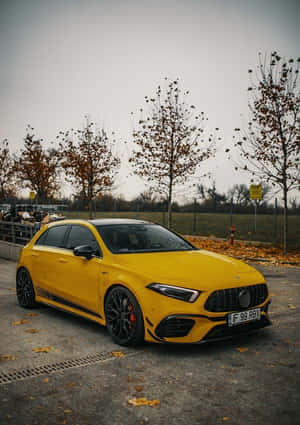 黃色 Mercedes-AMG A45 S 停放在人行道上，背景有落葉和光禿的樹木。 — 秋季 壁紙
