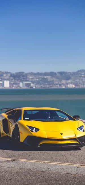 黃色藍寶堅尼Aventador停泊在海邊，遠處有城市。 — iPhone 14 Plus 壁紙
