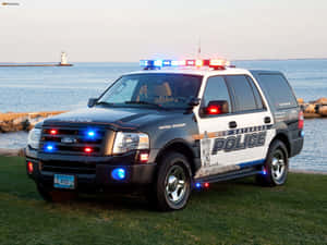 警用 SUV 閃爍著燈光，停在靠近水邊的地方，背景有燈塔。 — 福特 壁紙