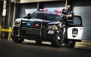 身穿黑色制服的警員從黑色道奇警車的打開車門探出身來，警車在小巷中閃著警燈。 — 警察 GIF 壁紙