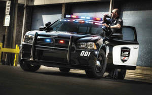 身穿黑色制服的警員從黑色道奇警車打開的車門探出身子，警車在小巷中閃著警燈。 — 汽車 壁紙