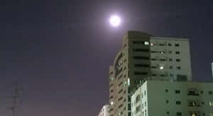 滿月在夜晚的城市建築上方發光，窗戶亮著燈。 — 美麗的滿月 壁紙